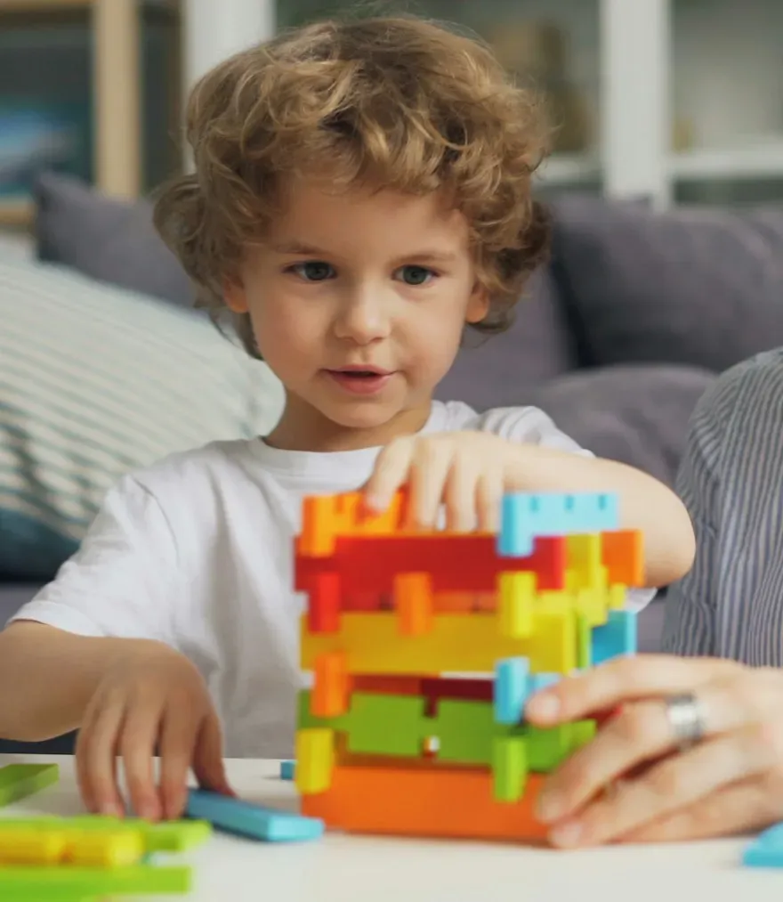 A young boy concentrating as he builds a colourful block tower, with an adult's hand steadying it beside him